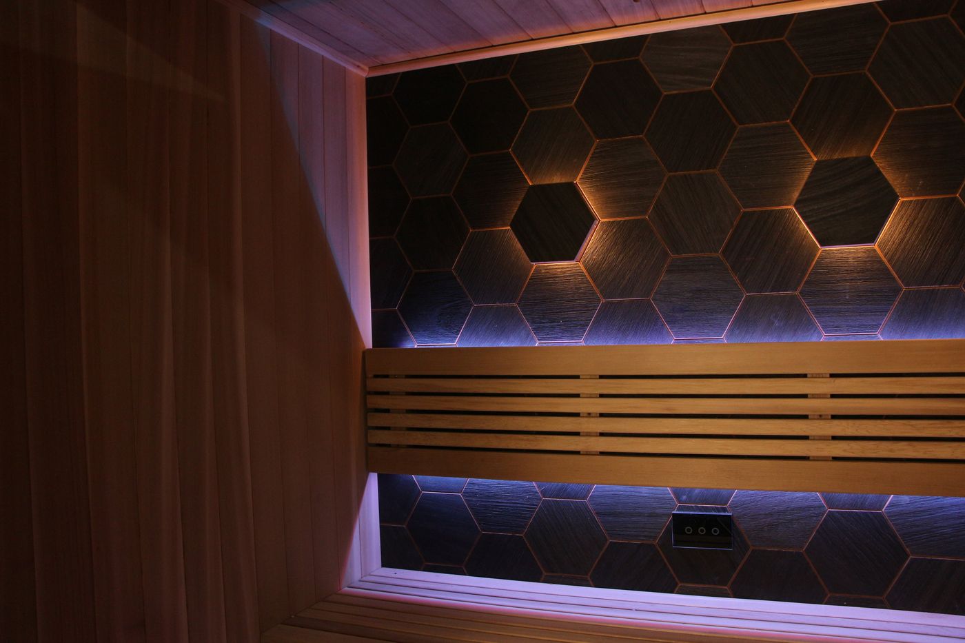 The interior of an empty modern sauna, featuring wooden benches and a back wall with dark, backlit hexagonal tiles.