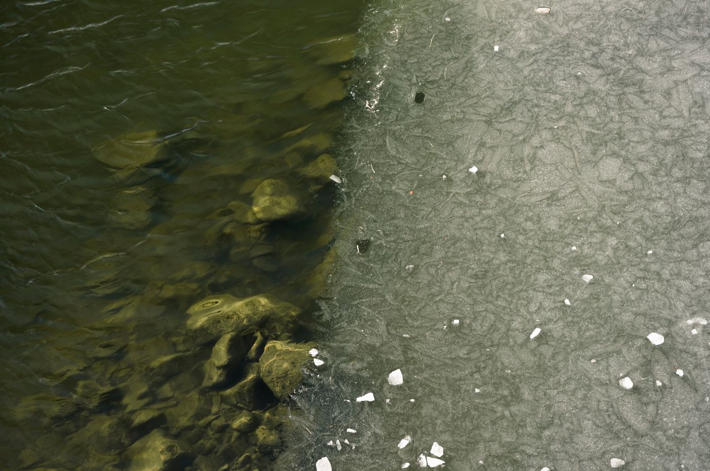 An overhead view of a body of water, with the left side showing clear water and rocks below, and the right side covered in a layer of ice.