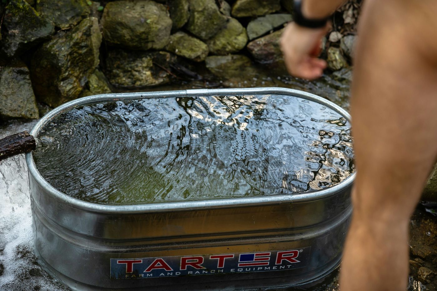A person stands next to a metal stock tank being filled with water outdoors in a rocky area.