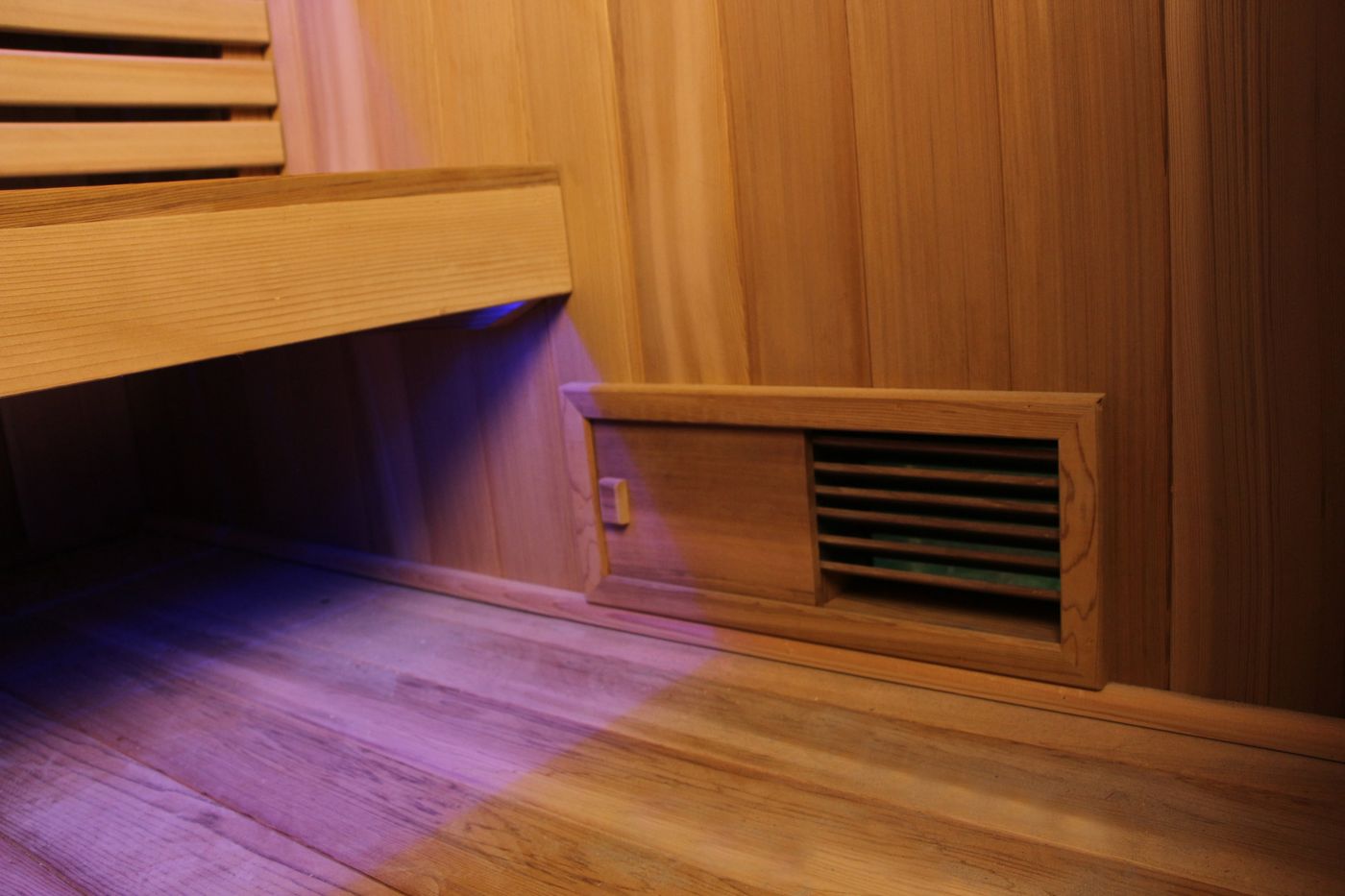 The interior of an empty wooden sauna, showing a bench, floor, and a vent, with purple light under the bench.
