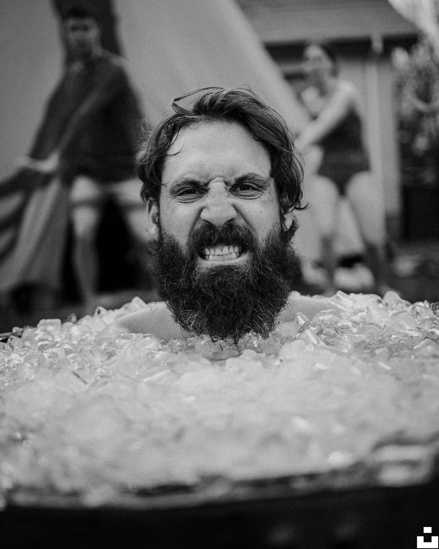 A black and white close-up photo of a bearded man grimacing while sitting in a tub full of ice.