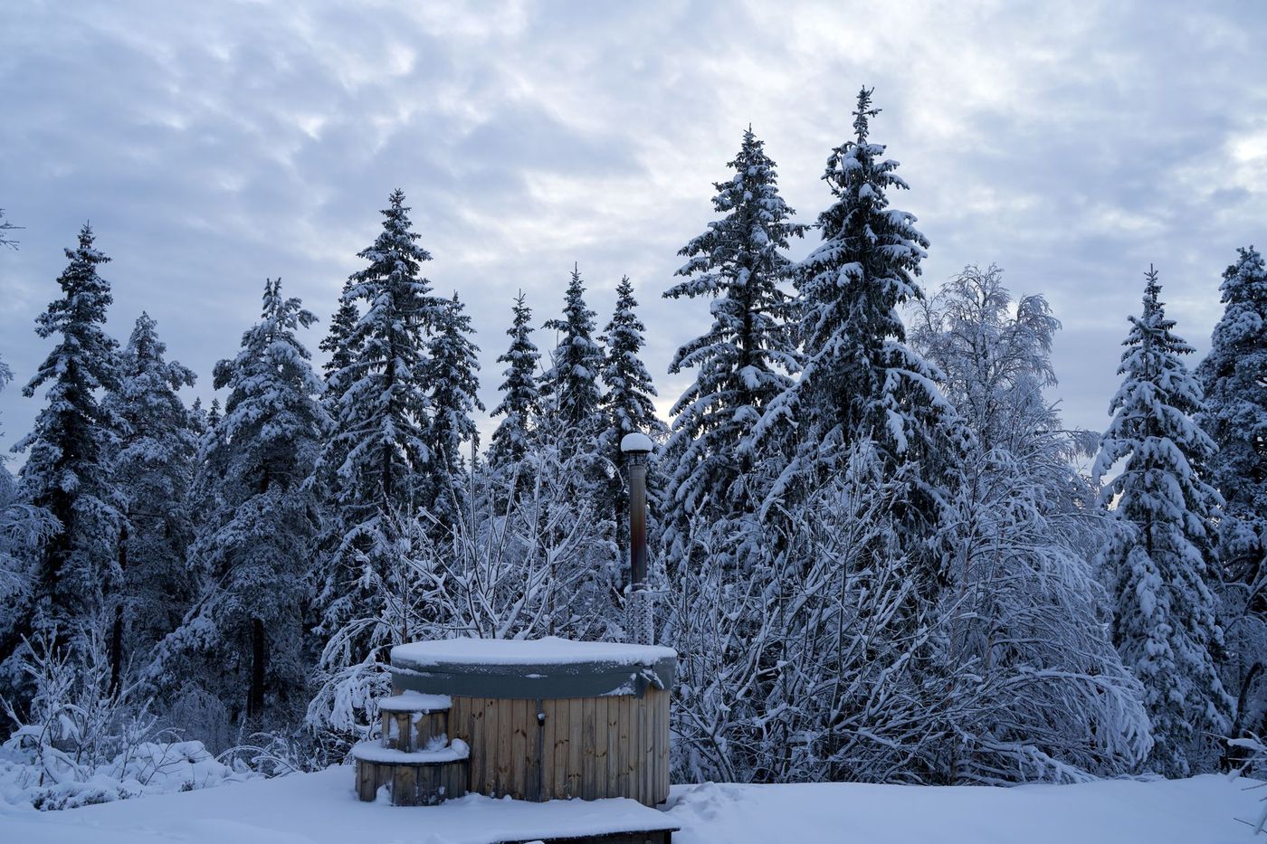 A wooden hot tub with a cover on it sits in a snowy landscape surrounded by snow-covered evergreen trees.