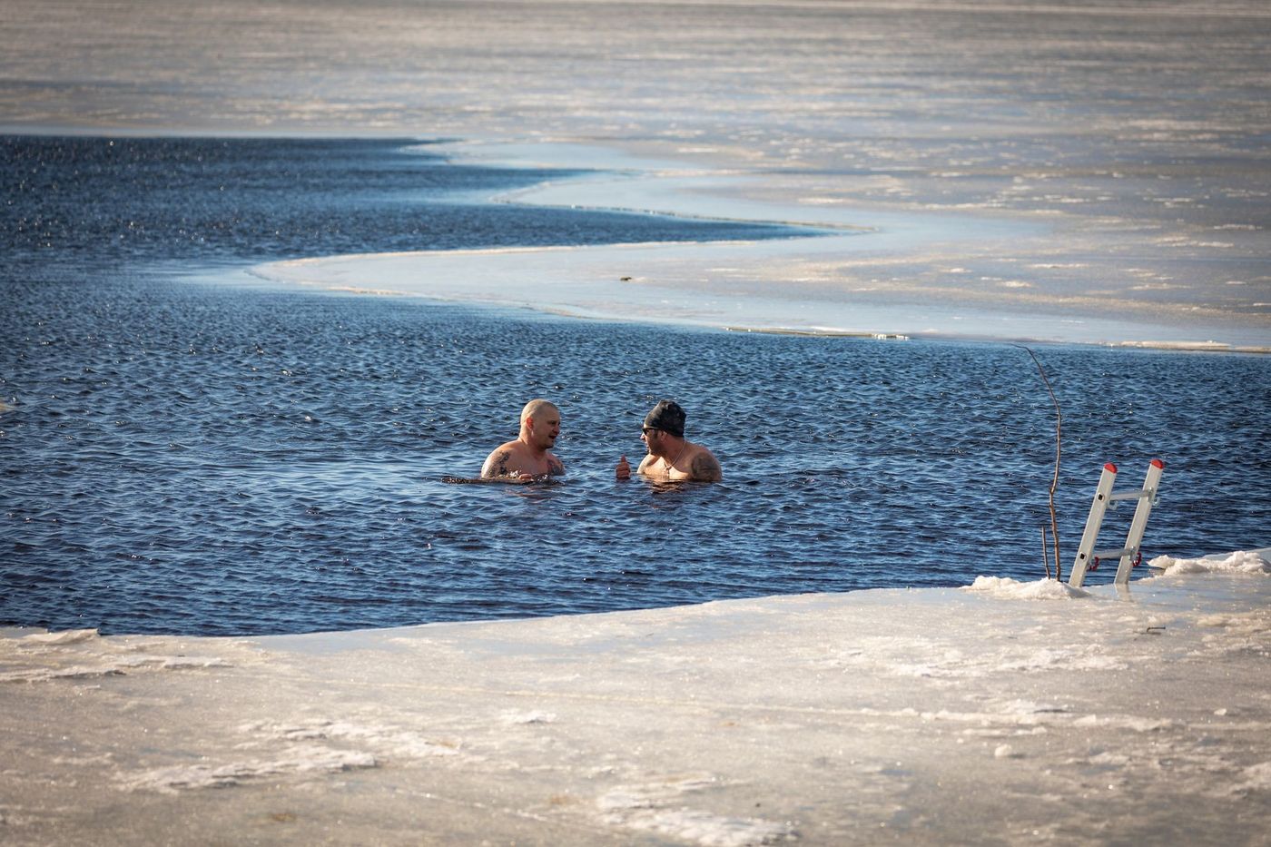 Two men are in the water of a partially frozen lake or river, talking to each other.