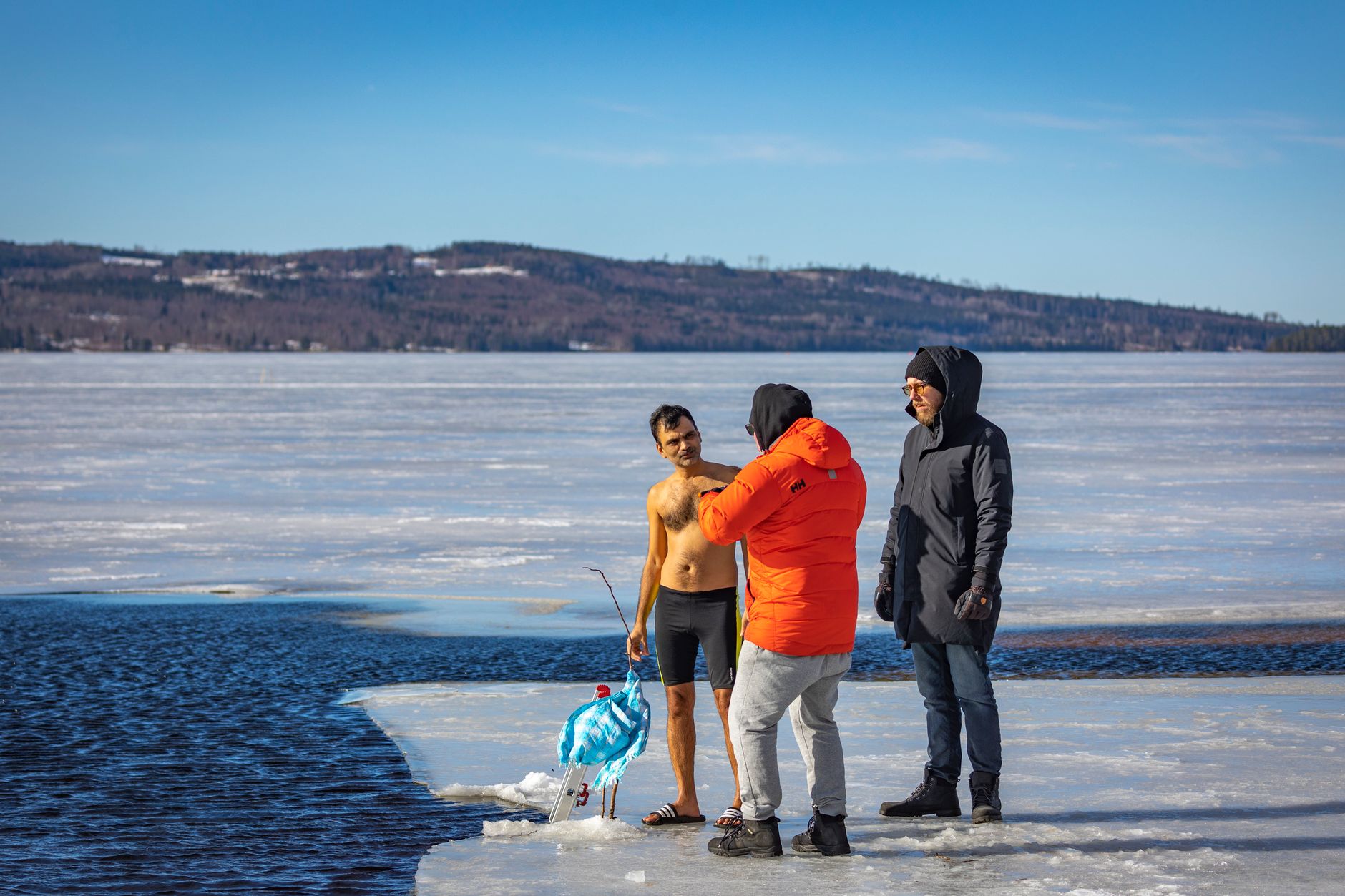 A man in swimming trunks stands on the ice at the edge of a frozen lake, talking to two other men who are dressed in winter coats.