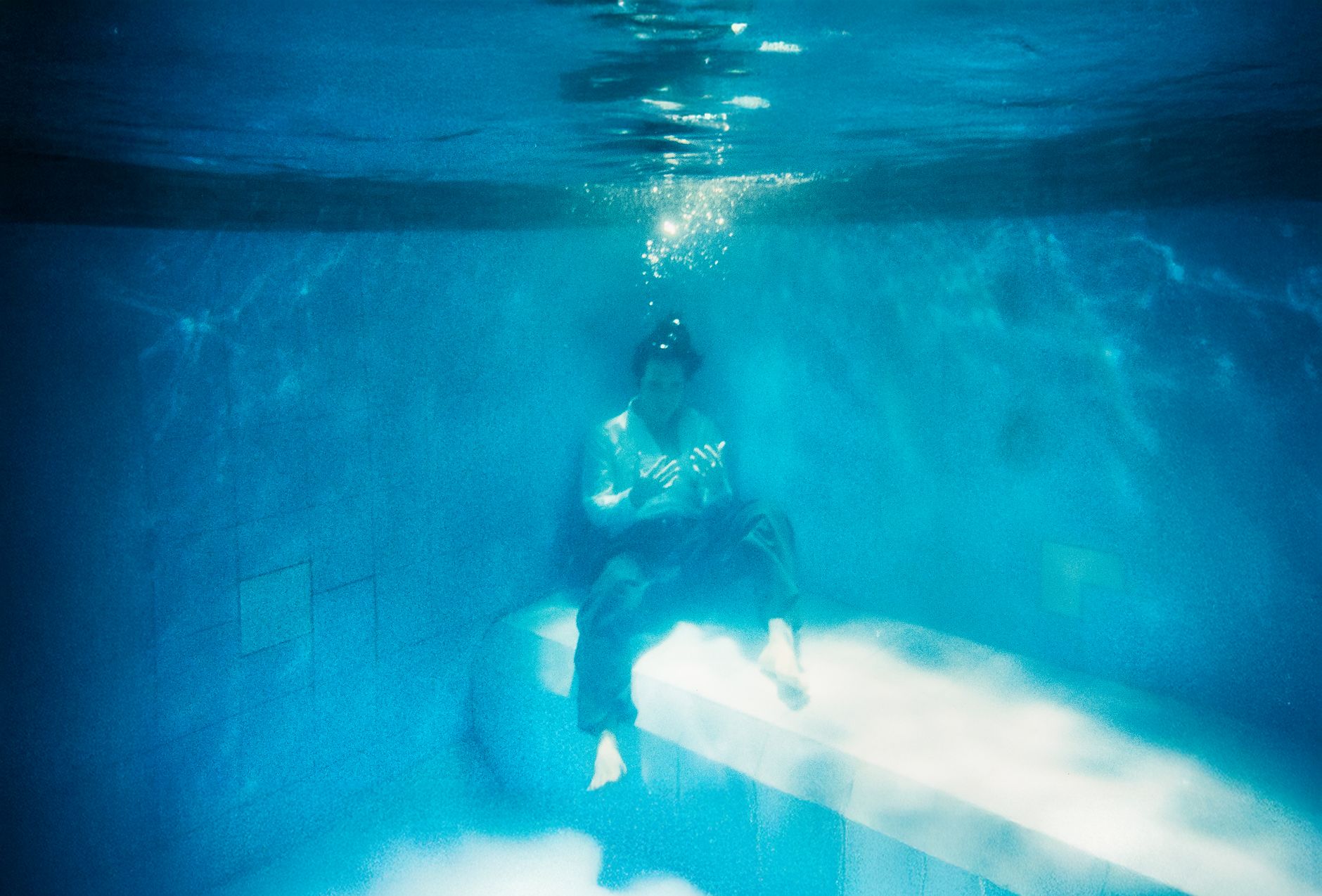 A fully clothed person sitting on a ledge completely underwater in a swimming pool.