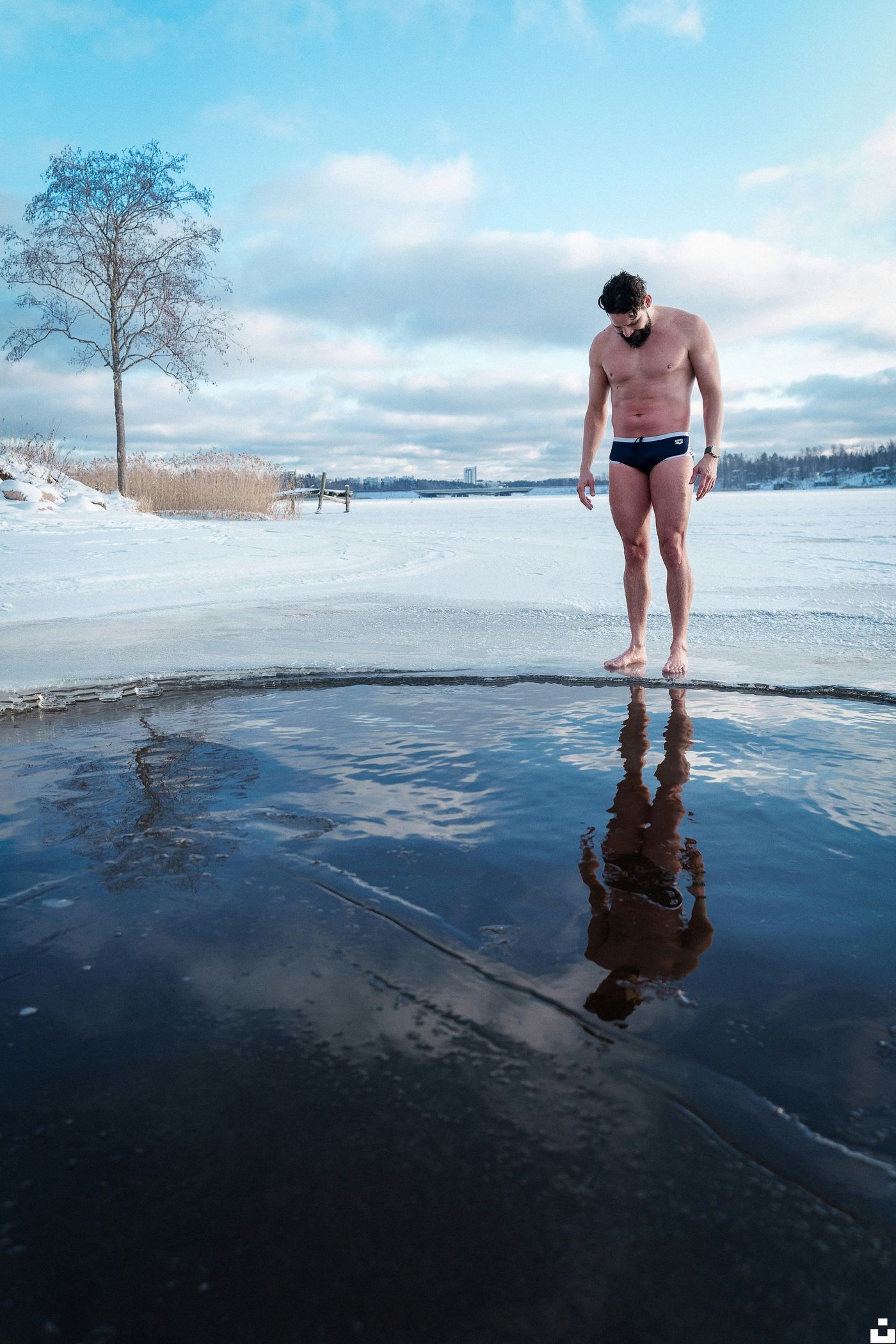 A man in swim briefs stands barefoot on the edge of a hole cut into a frozen lake, looking down at the water.