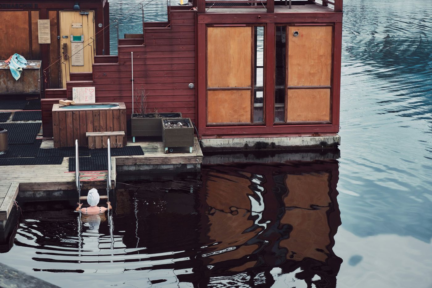 A woman with a towel on her head, seen from the back, is in the water holding a ladder attached to a wooden dock with a sauna.