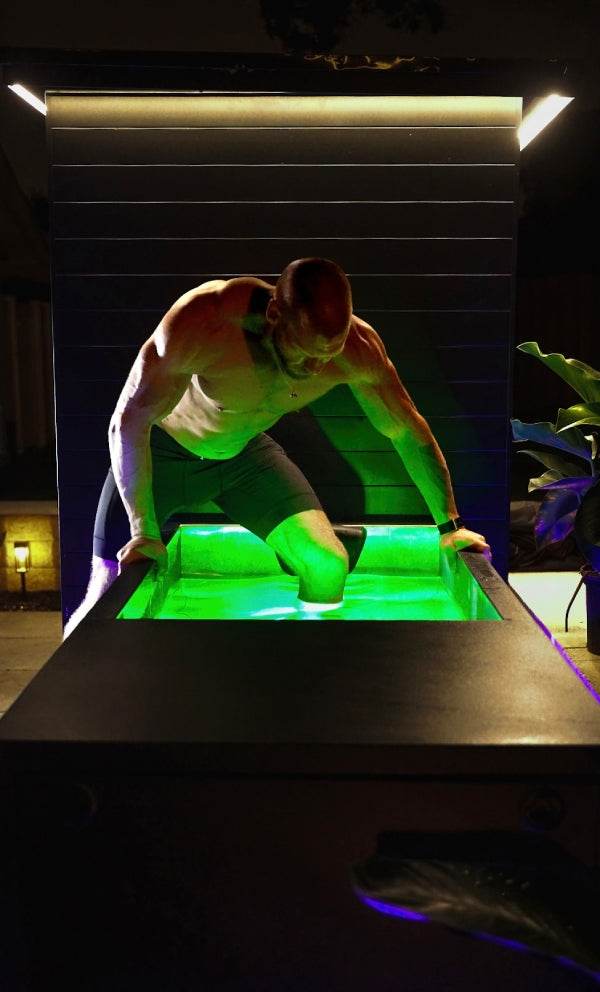 A muscular man in athletic shorts lowers himself into a modern cold plunge tub with glowing green water, outdoors at night.