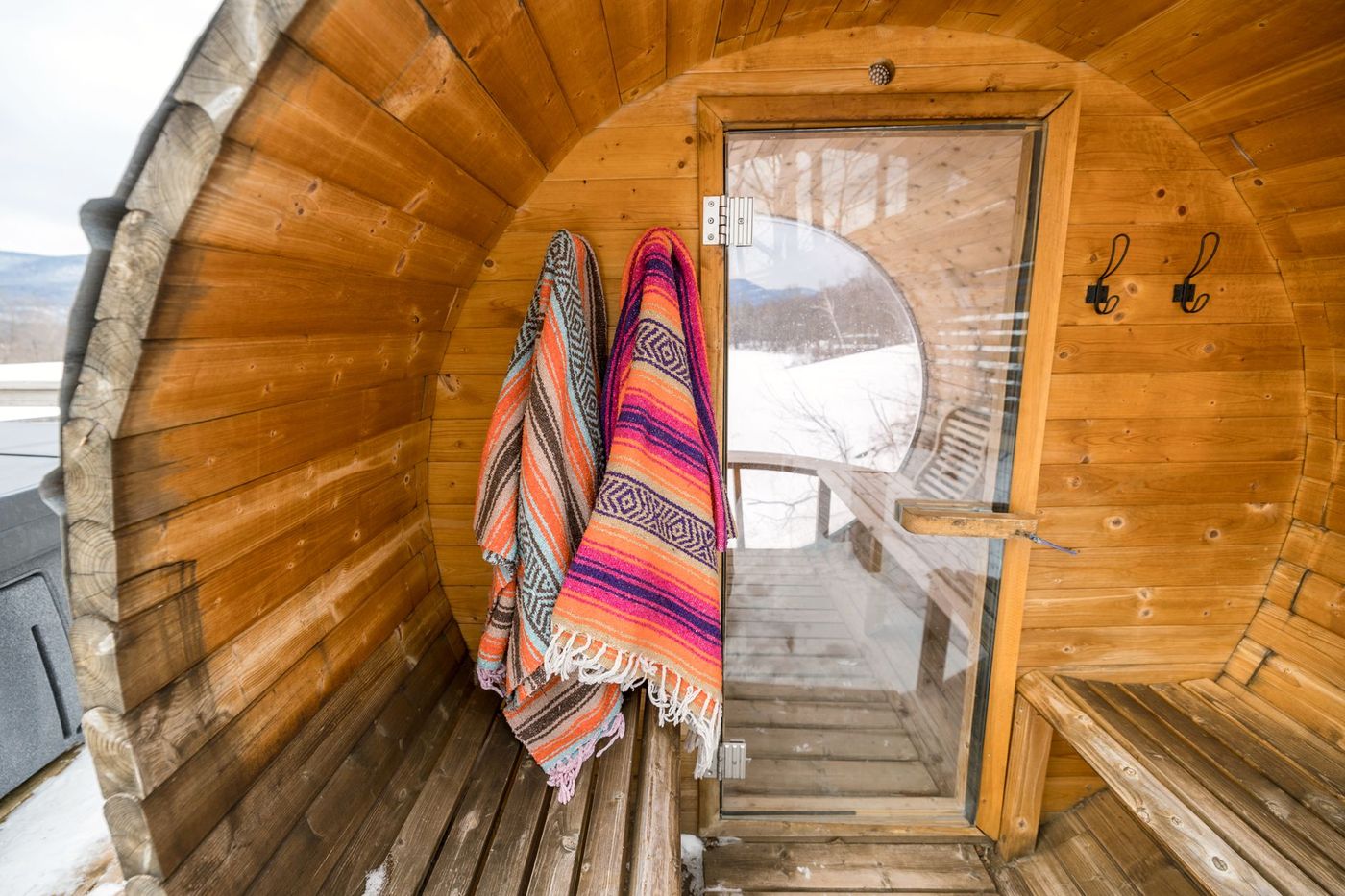 The interior of a wooden barrel sauna with two colorful woven blankets hanging on hooks, looking out a glass door to a snowy landscape.