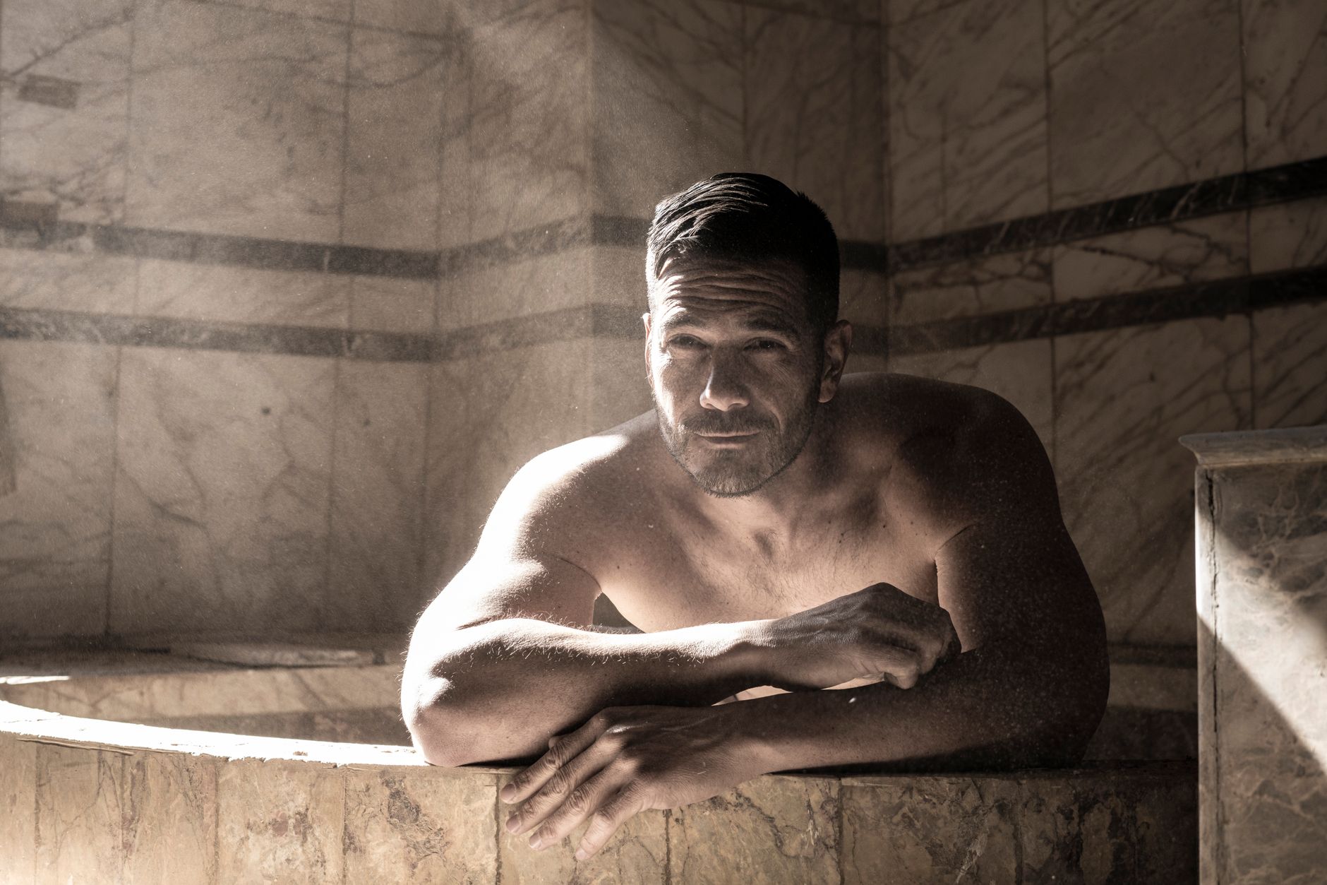A muscular man rests in a marble tub in what appears to be a bathhouse, leaning forward with his arms on the edge.