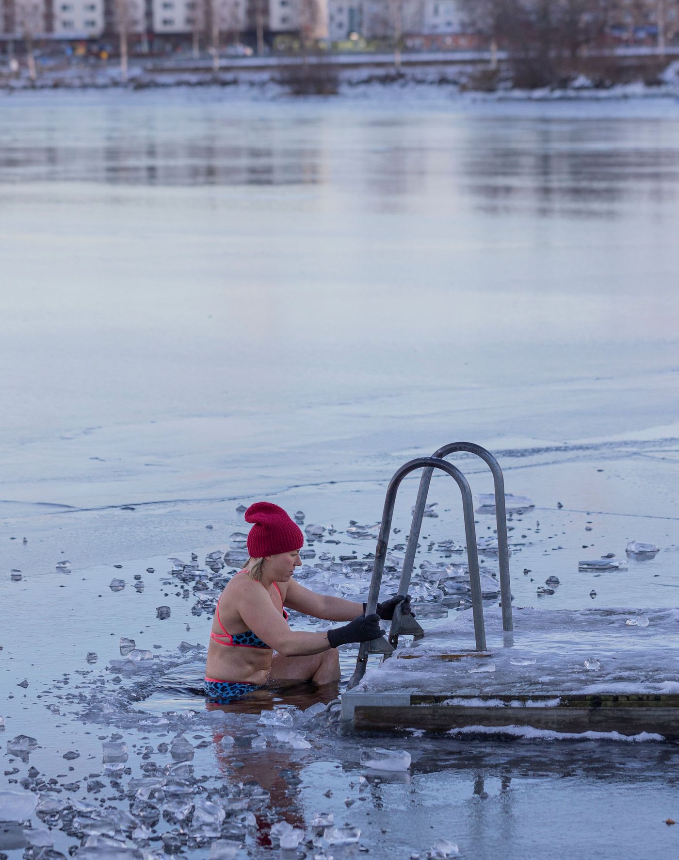 A person in a bikini and winter hat sits in a frozen lake or sea, holding a ladder attached to a dock.