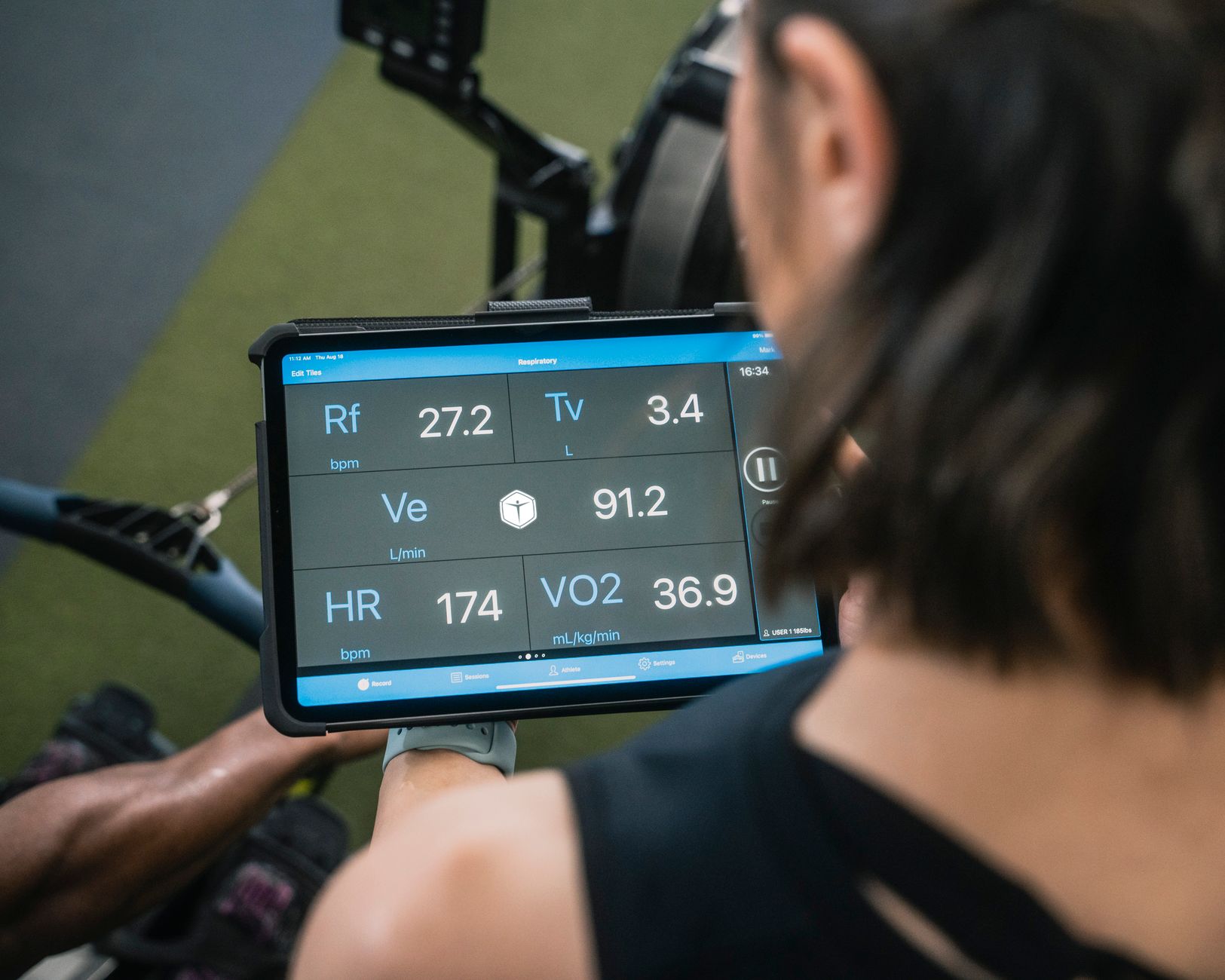 A person holds a tablet displaying biometric data like heart rate and VO2 max, likely monitoring someone exercising in a gym.