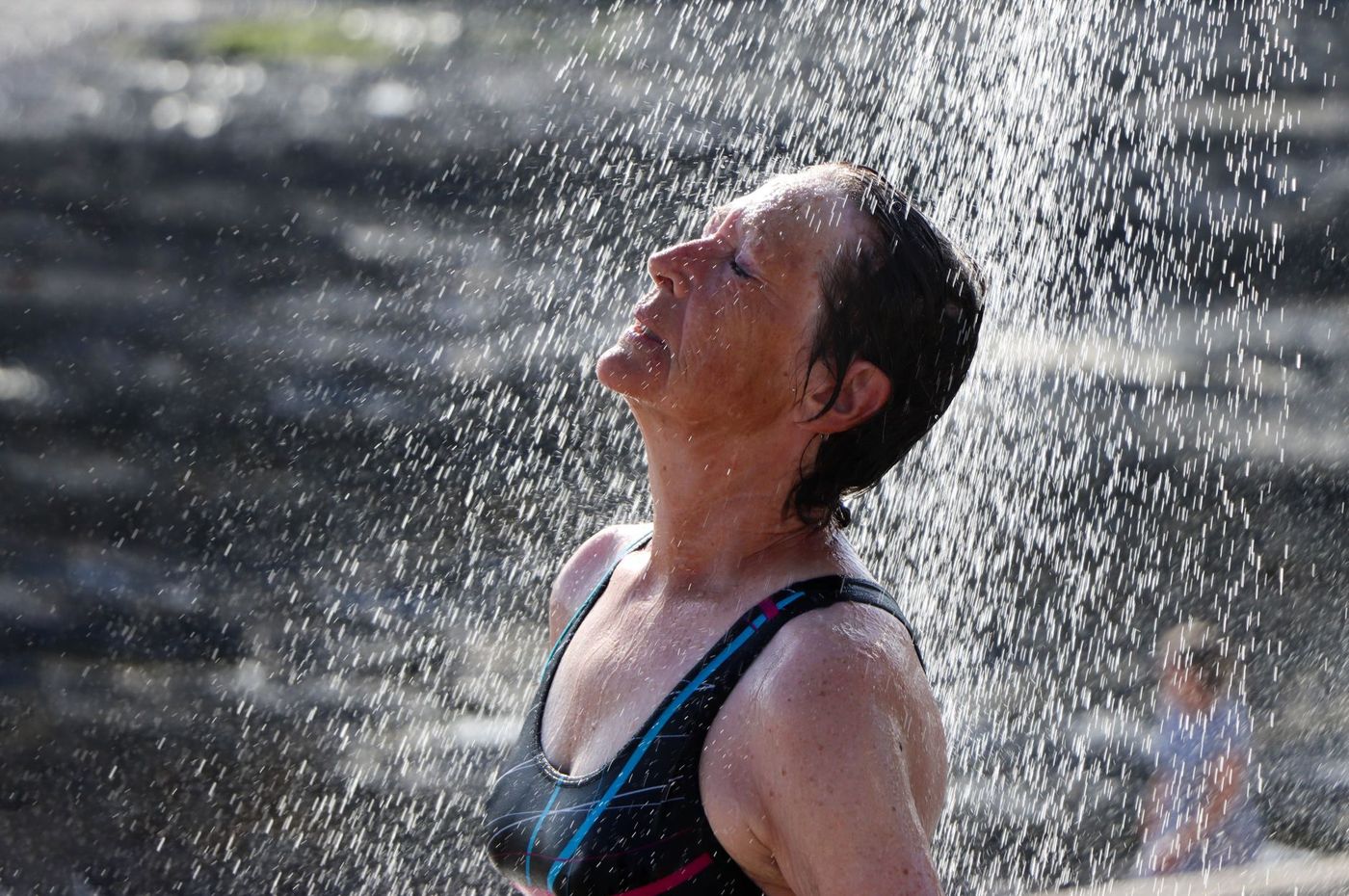 A woman in a swimsuit stands under an outdoor shower with her face tilted up towards the water.