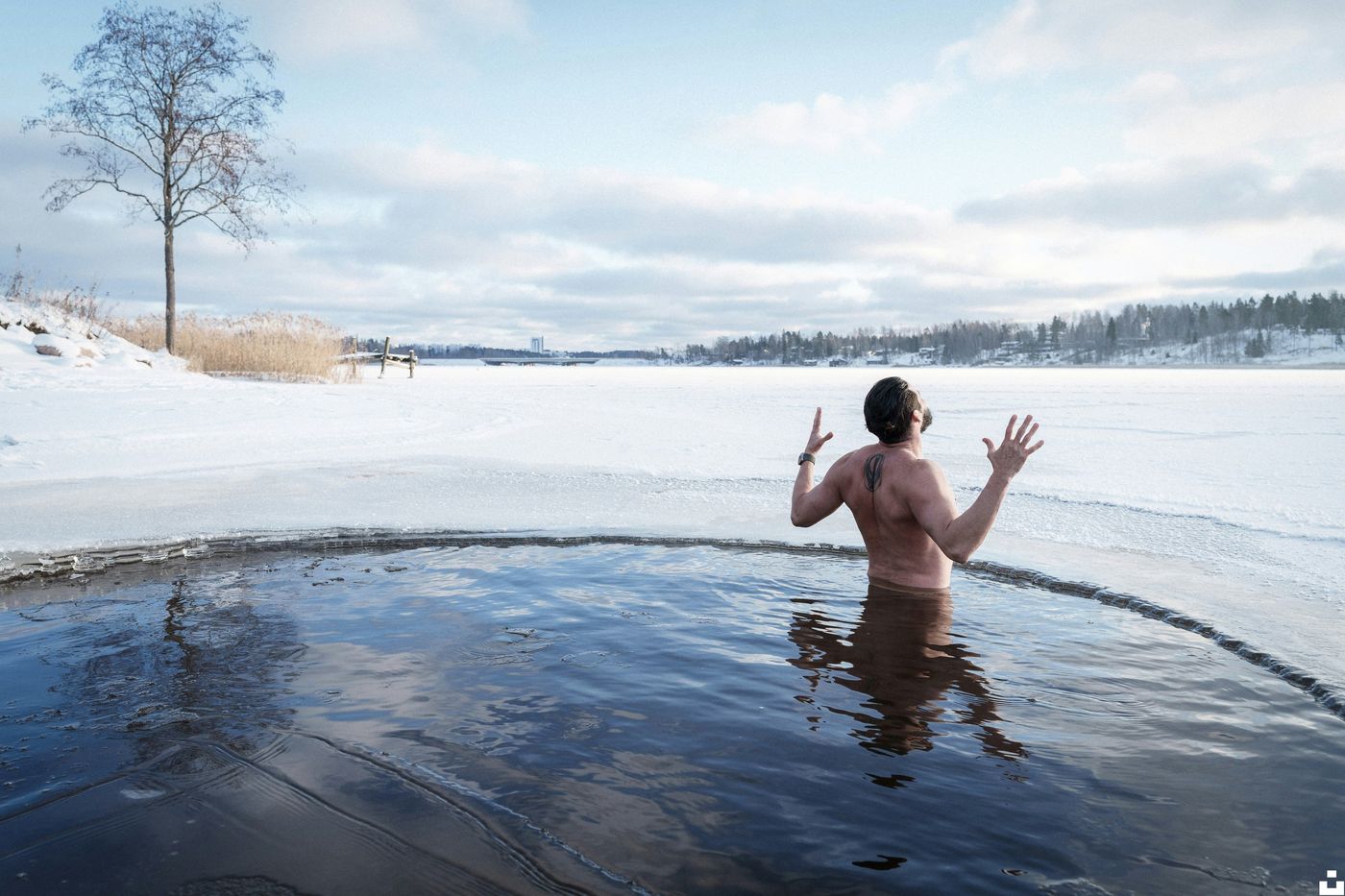 A man stands chest-deep in a hole cut into a frozen lake, with his arms raised in the air.