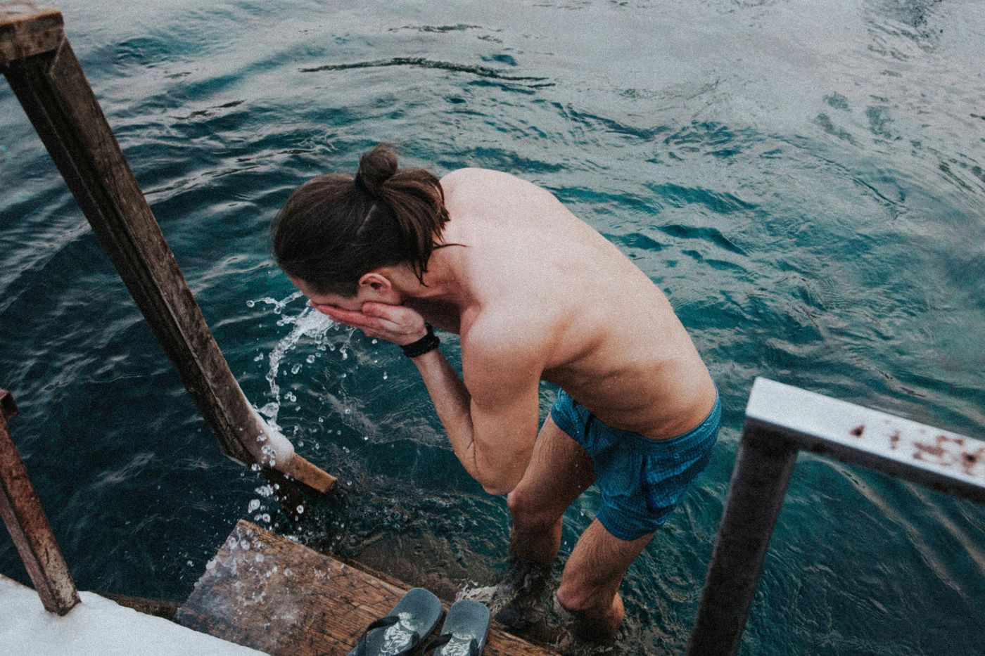A person with long hair in a bun stands on steps in a cold body of water, splashing water on their face.