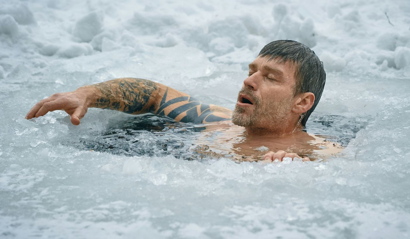 A man with a tattooed arm is submerged in a hole cut into a frozen body of water outdoors, with his eyes closed and mouth open.