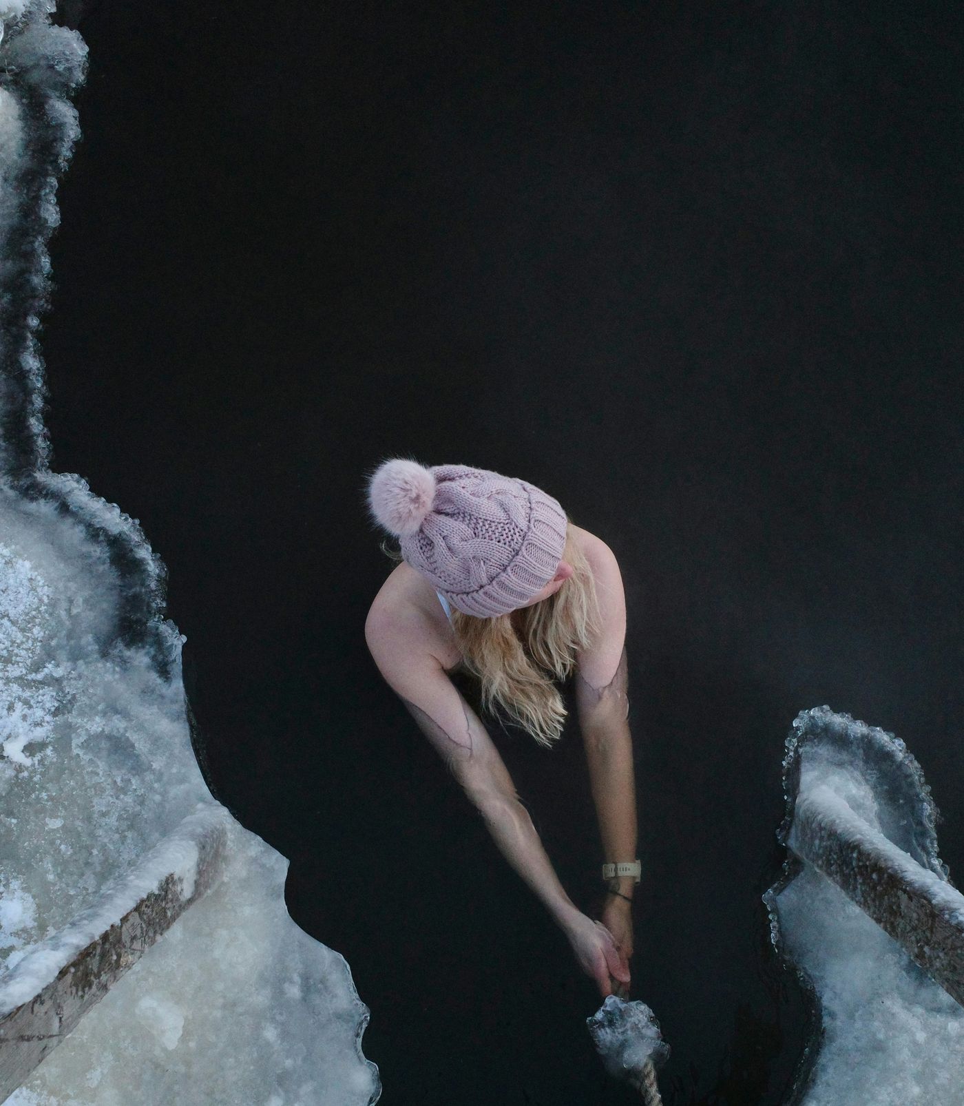 An overhead shot of a person wearing a knit hat in a hole cut into an icy body of water.