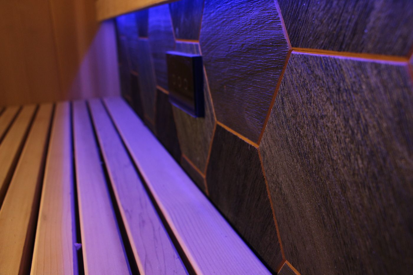 A close-up of a wooden bench inside a sauna with a dark, textured, hexagonal-tiled wall and blue ambient lighting.