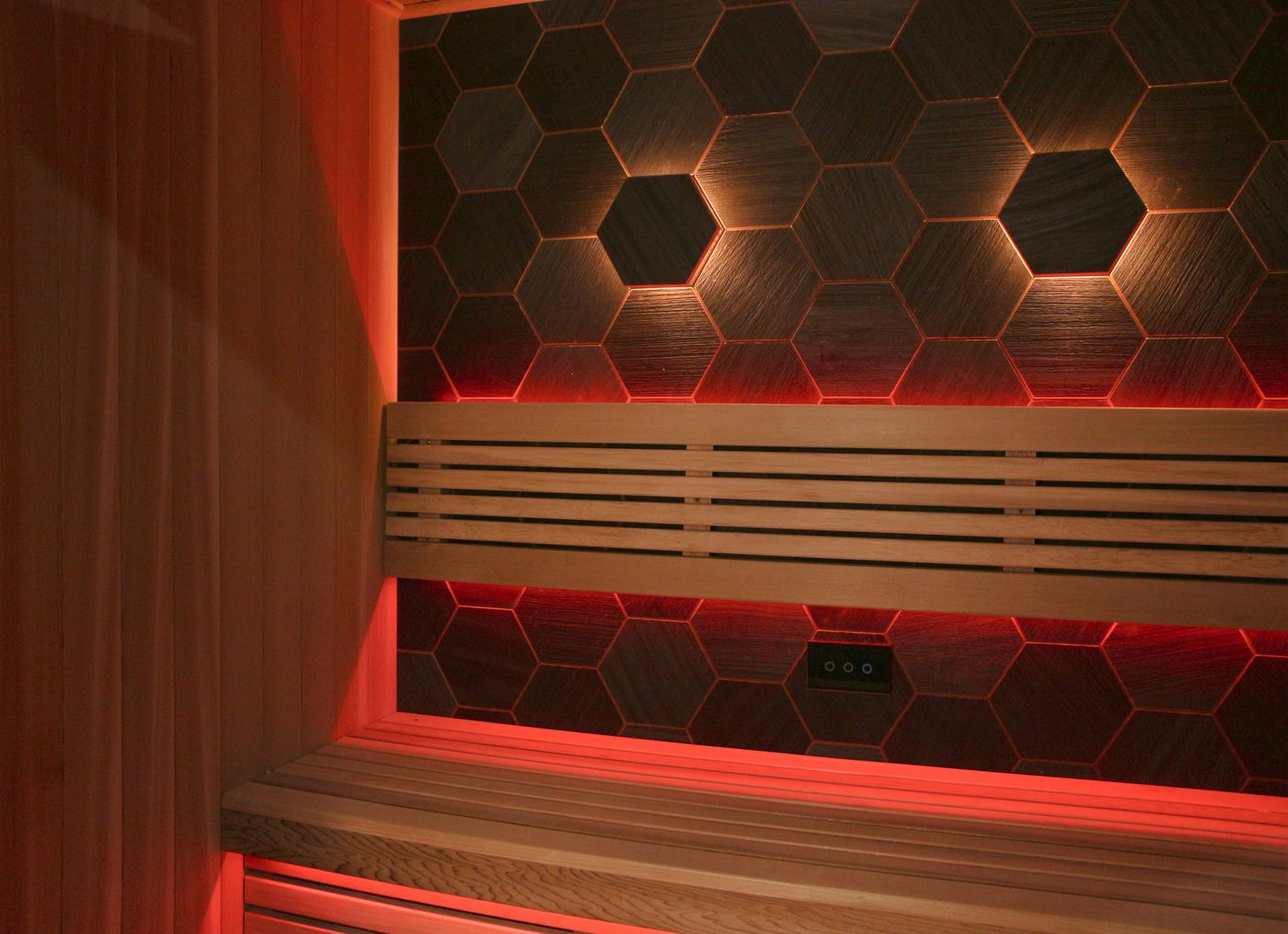 The interior of a modern, empty sauna with wooden benches, hexagonal tiles, and red and yellow mood lighting.