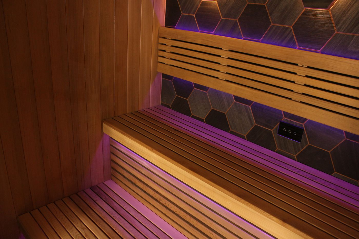 The empty interior of a modern sauna with wooden benches, hexagonal tiled walls, and purple mood lighting.