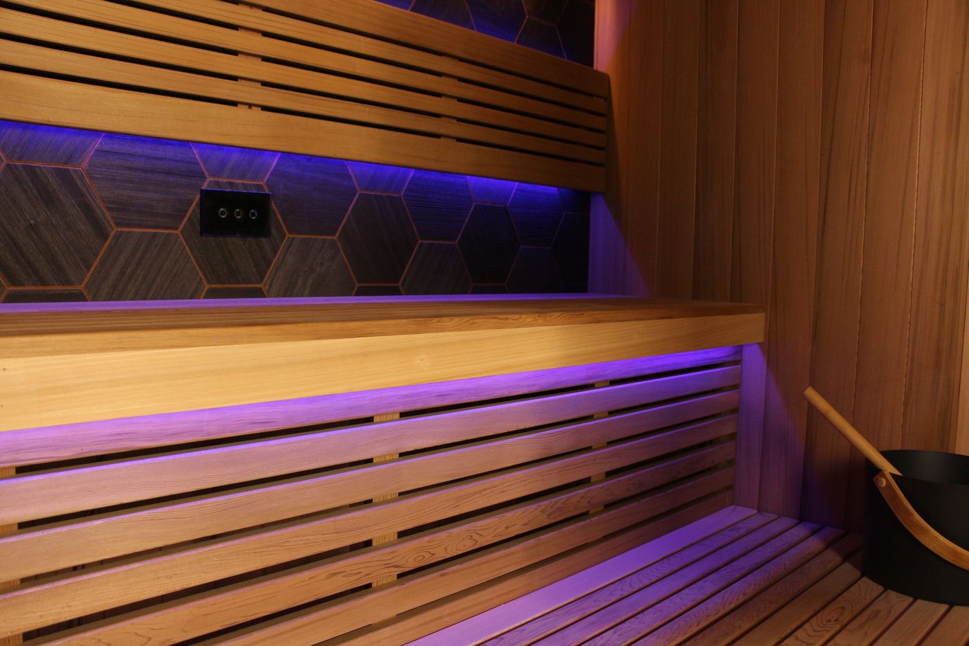 The interior of a modern wooden sauna with tiered benches, purple LED lighting, and a bucket with a ladle in the corner.