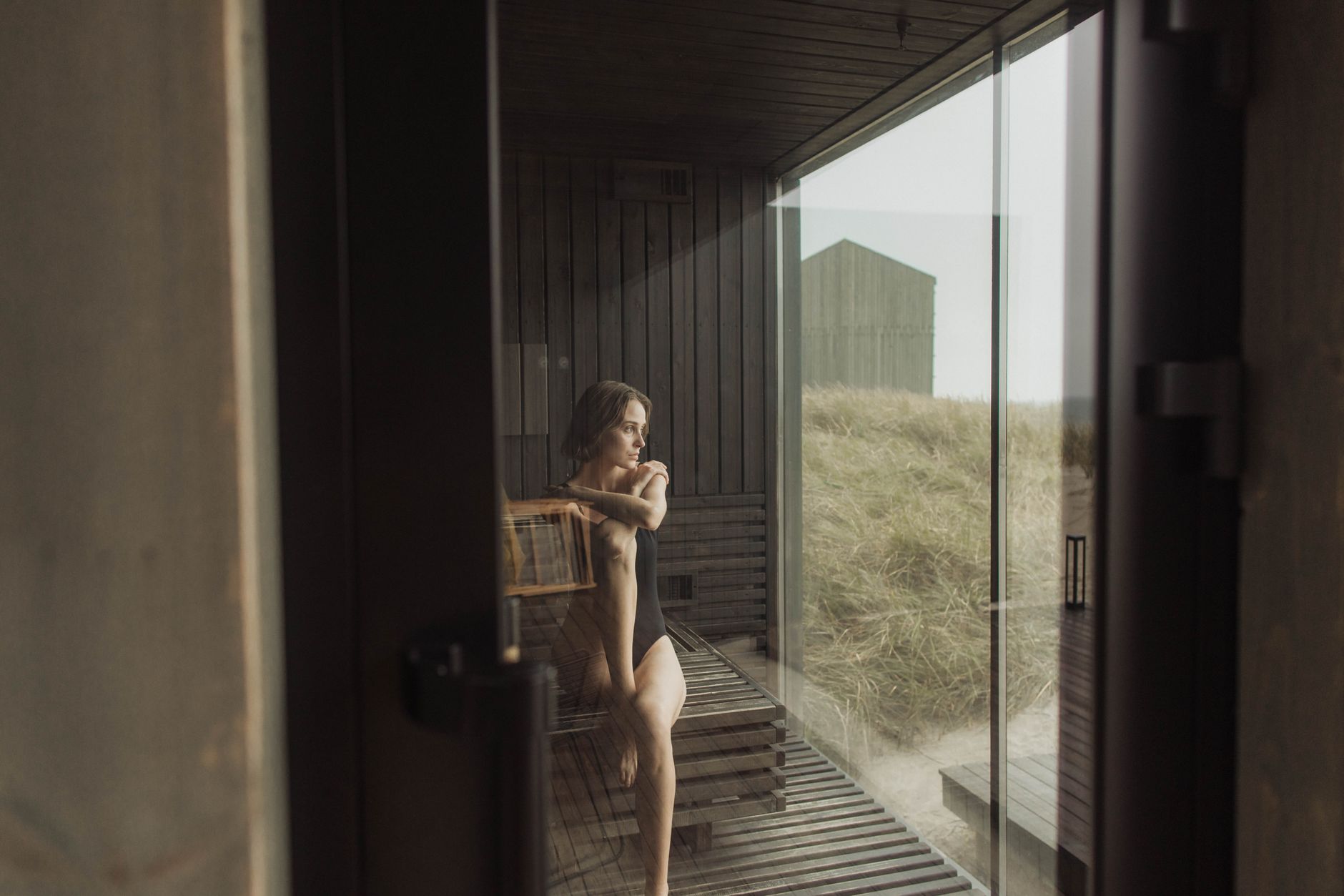 Woman in a wooden infrared sauna facing a stainless steel cold plunge tub on a sunny patio