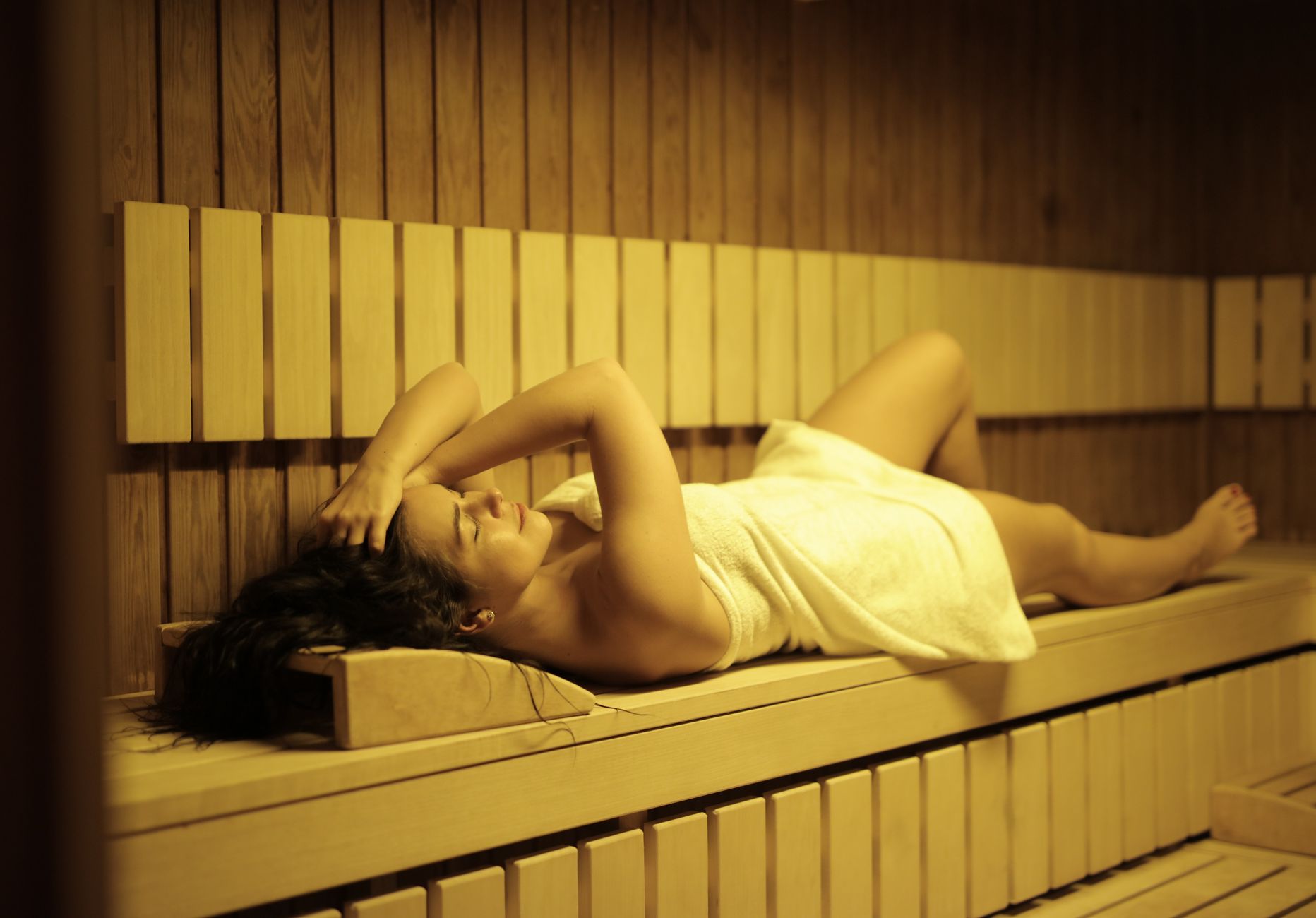 A woman wrapped in a towel is lying down and relaxing on a wooden bench inside a sauna.