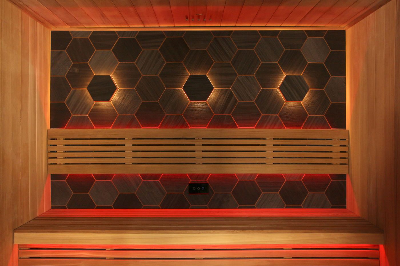 The interior of an empty wooden sauna with two levels of benches, a back wall with dark hexagonal tiles, and red and white accent lighting.