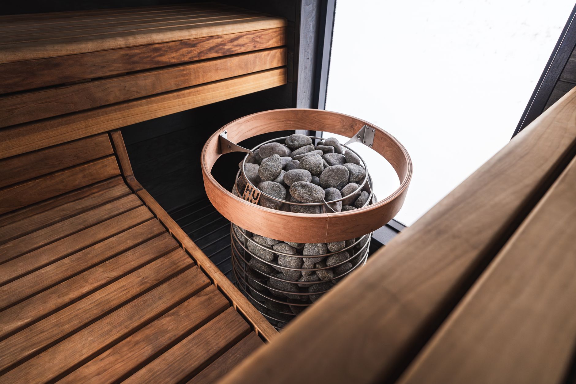 The interior of a modern wooden sauna, focusing on a cylindrical heater filled with stones next to the benches.