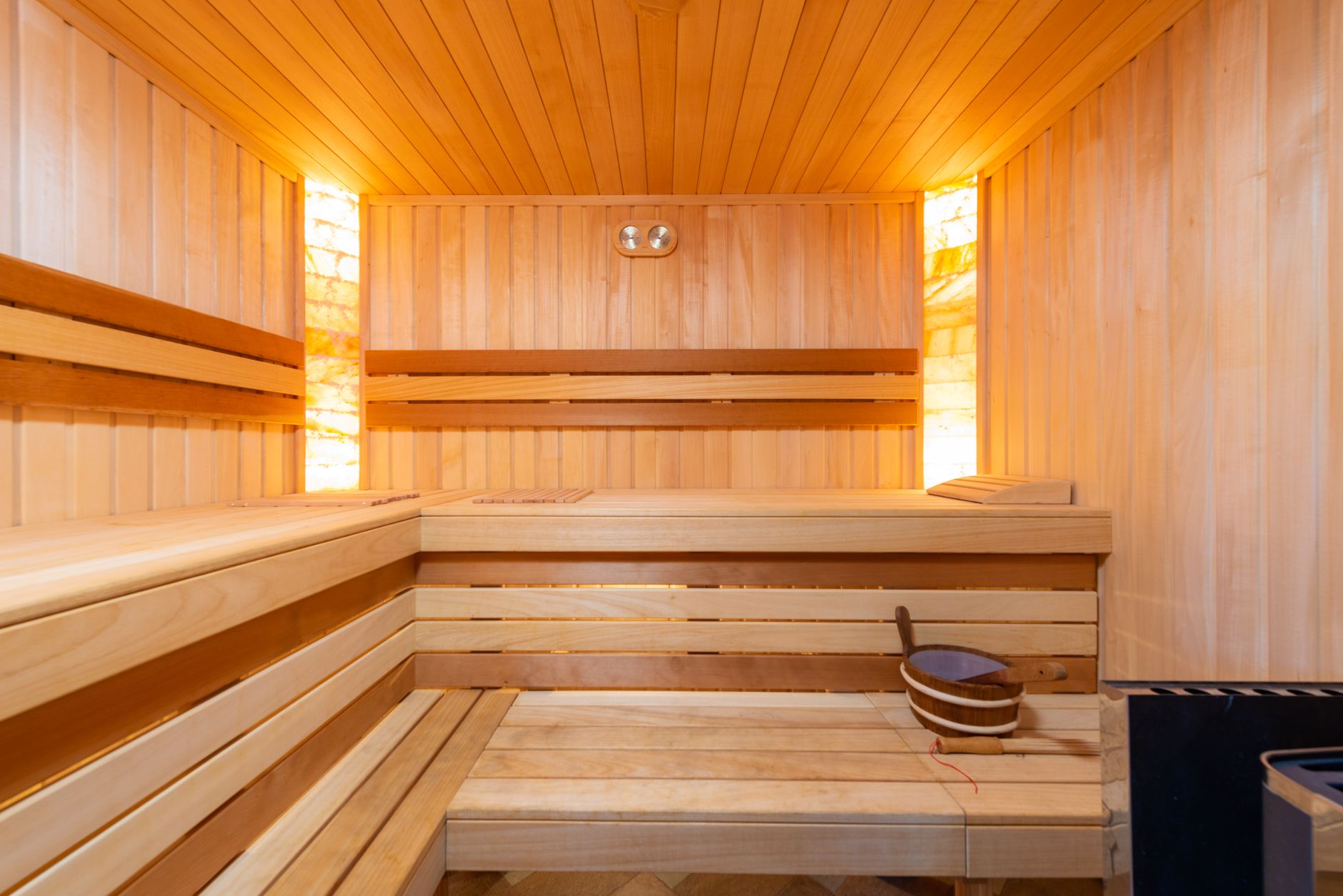The interior of a modern wooden sauna with tiered benches, warm lighting, and a bucket with a ladle.