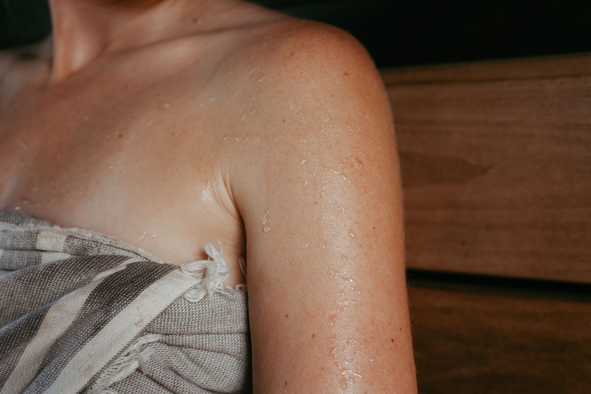 A close-up of a person's sweaty shoulder and arm, wrapped in a striped towel, inside what appears to be a wooden sauna.