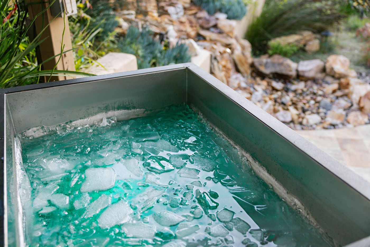 A close-up of a metal ice bath tub filled with turquoise water and ice cubes, set in an outdoor, landscaped area.